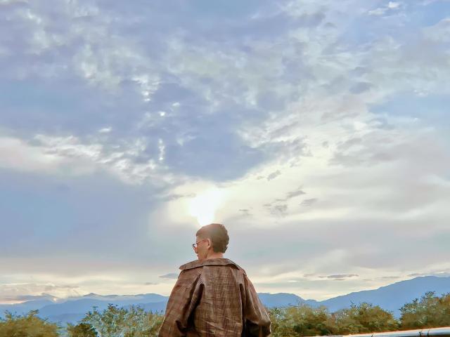 SantiagoDairyFarmer with short brown hair, wearing glasses and a brown plaid coat, standing with back to camera, looking at a cloudy, sunlit sky over mountains.