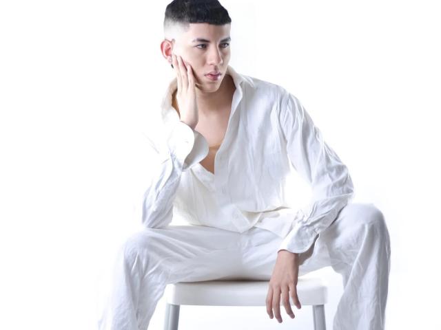 SantiagoDairyFarmer, with short black hair, sits on a white stool, wearing a loose white shirt and pants, hand on cheek, looking thoughtful, in a bright studio shot.