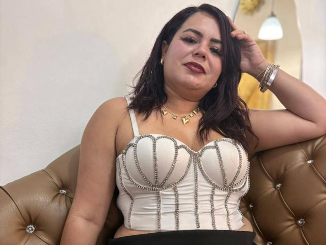 MayaMalik with dark hair, wearing a white, studded, strapless top, showing her big, shiny tits, sitting on a tan, tufted leather couch, hand in hair, gold necklace.