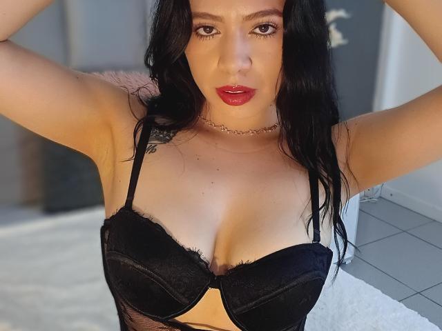 Linda Mancini met zwarte beha, grote borsten, lichte huid, lang zwart haar, rode lippen, choker, armen omhoog, kijkt naar de camera, witte betegelde vloer, zacht licht..