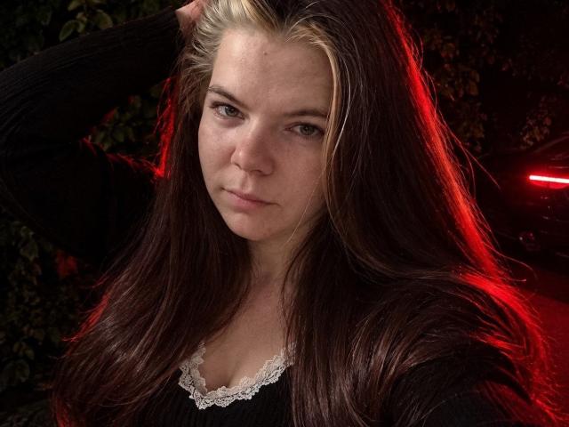 RonnieFarr met lang bruin haar, een blonde pluk, hand in haar haar, serieuze uitdrukking, draagt een zwarte top met kantrand, rood licht op de achtergrond. Borsten licht zichtbaar onder de kant..