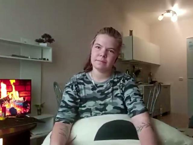 Ronnie Farr mit langem, braunem Haar, in einem Tarnhemd, sitzt auf einem Sofa, hat Tätowierungen an den Armen und blickt in die Kamera. Im Hintergrund ist ein Fernseher mit einem Bild von Feuer zu sehen, der Raum ist weiß und es gibt eine Deckenleuchte..