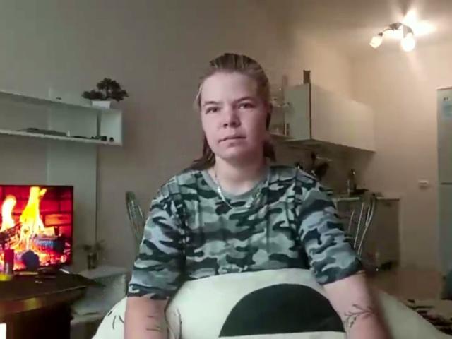 RonnieFarr with long brown hair in a camo shirt, sitting on a couch, looking at the camera with a neutral expression, tattoos on arms, TV with fire on left.
