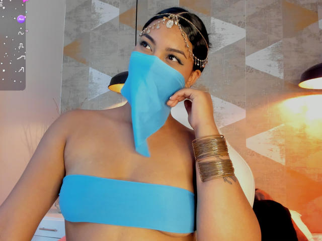 LalilaBrunette, peau bronzée, cheveux noirs, portant un masque en tissu bleu, une chaîne dorée pour la tête et des bracelets dorés, montrant ses seins dans un haut bleu sans bretelles..