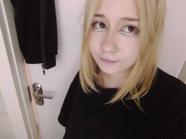 AnnieLeonhart69 has blonde hair, light skin, and brown eyes. She's wearing a black shirt, standing near a white door with a silver handle and black hanging item.