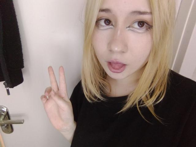 AnnieLeonhart69 with blonde hair, fair skin, and pink lips, making a peace sign with her right hand, wearing a black shirt, and sticking her tongue out.