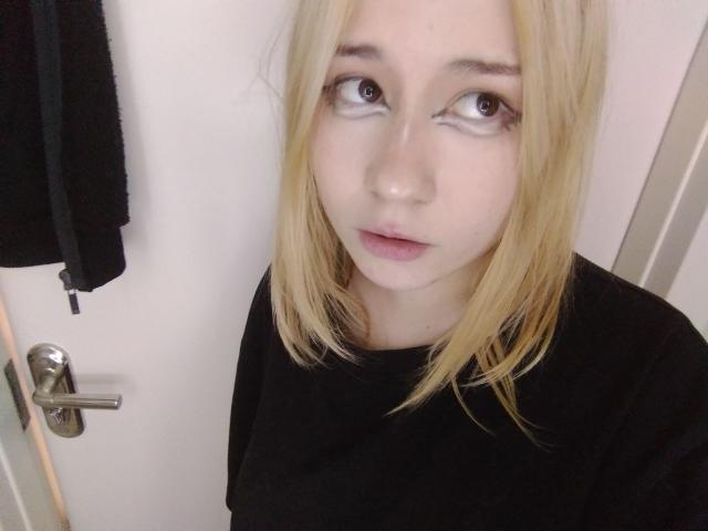 AnnieLeonhart69 has blonde hair, pale skin, and brown eyes, wearing a black shirt, looking up with a slightly parted mouth, standing in front of a white door.