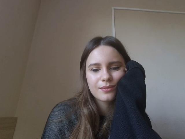 NellaKoczur ha lunghi capelli castani, eyeliner nero e un leggero sorriso, indossa un maglione nero e la sua mano destra tocca la testa. Il suo viso è al centro della foto..