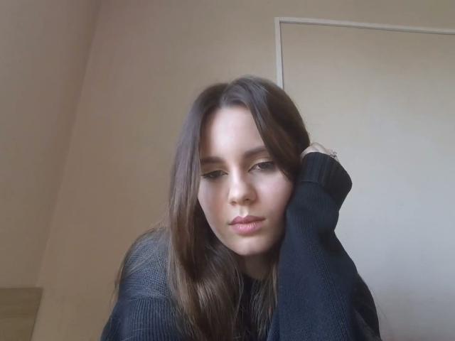 NellaKoczur with long brown hair, black sweater, hand on head, looking down with a slightly pouty expression, soft natural light, slight shadow on face.