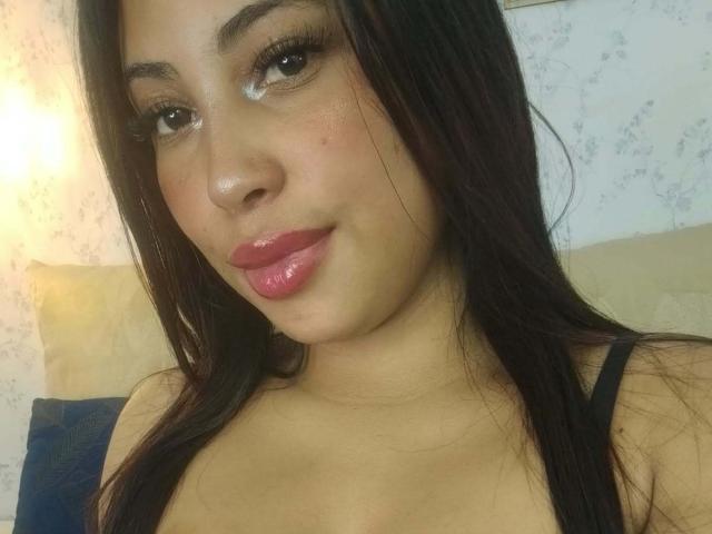 AndreaAleman has shiny pink lipstick, long dark hair, and is wearing a black spaghetti strap top, showing a little cleavage. She has a cute, slightly chubby face.