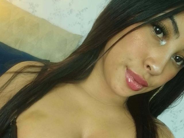 AndreaAleman has shiny brown eyes, pink glossy lips, and long dark hair. She's smiling with a slight nose bump, wearing a black top, showing her tits.