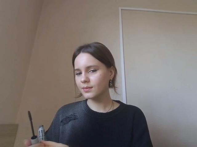 Nella Koczur, à la peau claire et aux cheveux bruns, portant une chemise noire texturée, tenant une petite bouteille, se tient debout dans une pièce avec une porte blanche en arrière-plan..