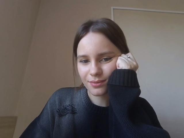 NellaKoczur with dark brown hair, black eyeliner, and a subtle smile, wearing a black sweater with a small embroidered design, resting her chin on her hand.