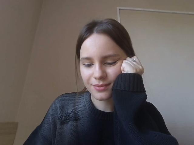 NellaKoczur, with straight brown hair, black eye makeup, and a black knit sweater, rests her cheek on her hand, smiling softly, showing a ring on her finger.