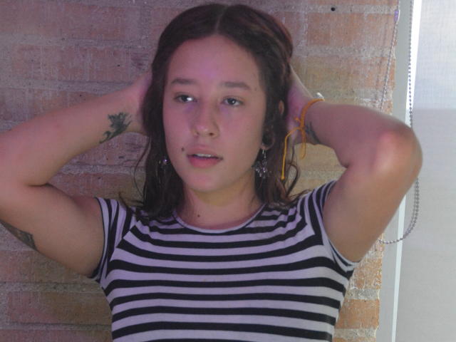 JuliethCourtouis with arms up, wearing a striped black and white shirt, has a small tattoo on her left arm and orange earbuds, looking slightly bored.