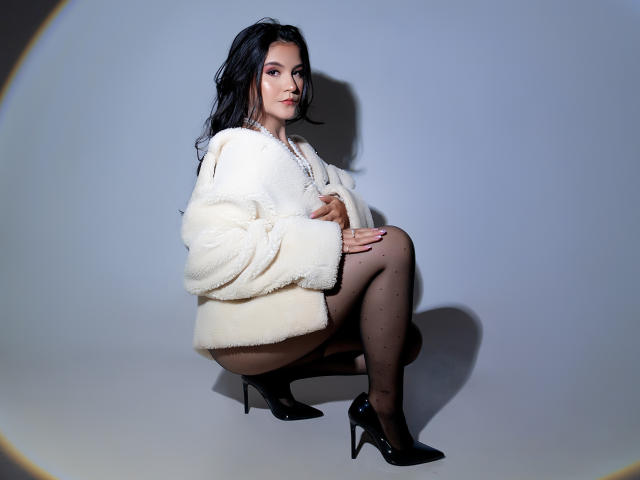 VelisseNoir está agachada, usando saltos altos pretos, um casaco branco fofo, collant preto com bolinhas e um colar de pérolas, com o bumbum ligeiramente saliente, e o cabelo escuro caindo..