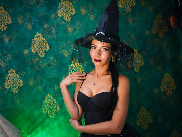 EvelinLuna con trucco scuro, cappello da strega nero e corpetto nero senza spalline, che mostra il seno, con una mano vicino al viso, luce verde sullo sfondo..