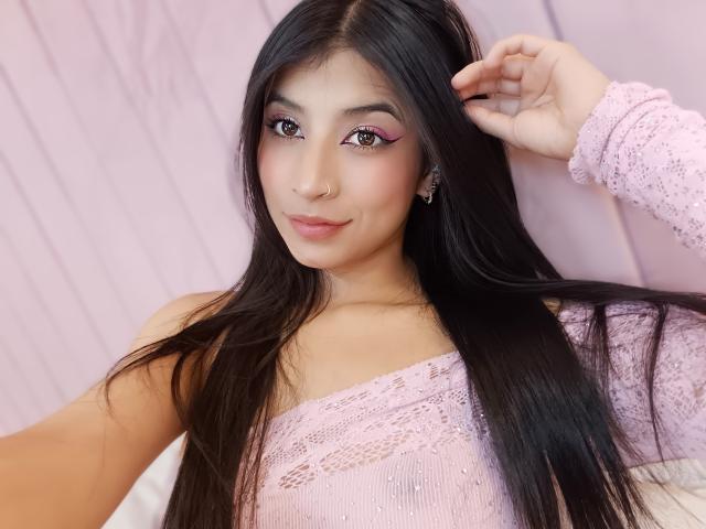 Naia Sanders, avec ses longs cheveux noirs, son eye-liner rose, son anneau de nez et son haut en dentelle rose, sourit et tient ses cheveux. Elle porte un anneau de nez et affiche un sourire charmant..