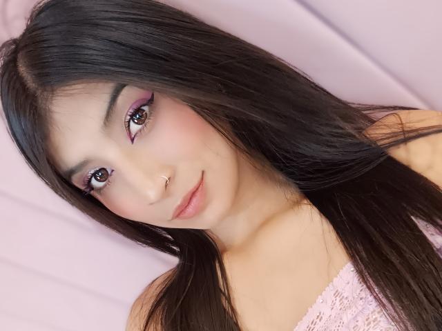 Naia Sanders, avec ses longs cheveux noirs, son eye-liner violet et son rouge à lèvres rose, porte un haut en dentelle rose, a un piercing au nez et affiche un léger sourire. Ses yeux sont marron et elle est jolie..