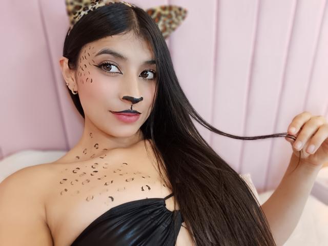 Naia Sanders, avec ses longs cheveux noirs, portant un bandeau léopard, un haut noir sans bretelles et du maquillage noir sur le visage avec des points éparpillés. Elle se tient les cheveux, mettant en valeur de jolis boucles d'oreilles..