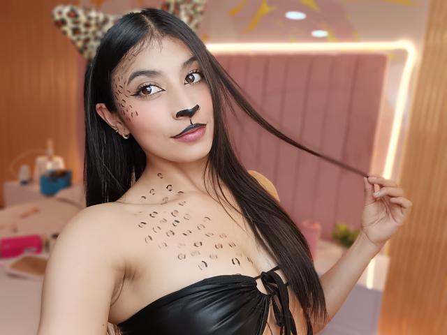 NaiaSanders with black lipstick, dark eye makeup, leopard print face paint, holding long black hair, wearing a black leather top with a ring, in a brightly lit room.