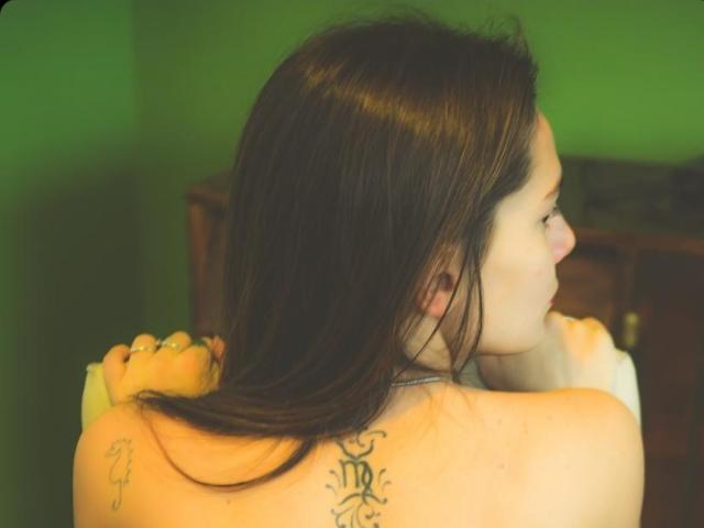 RomiSweety's bare back with a black tattoo of a symbol between her shoulders, brown hair, arms crossed, green background, wooden cabinet in the blurred background.