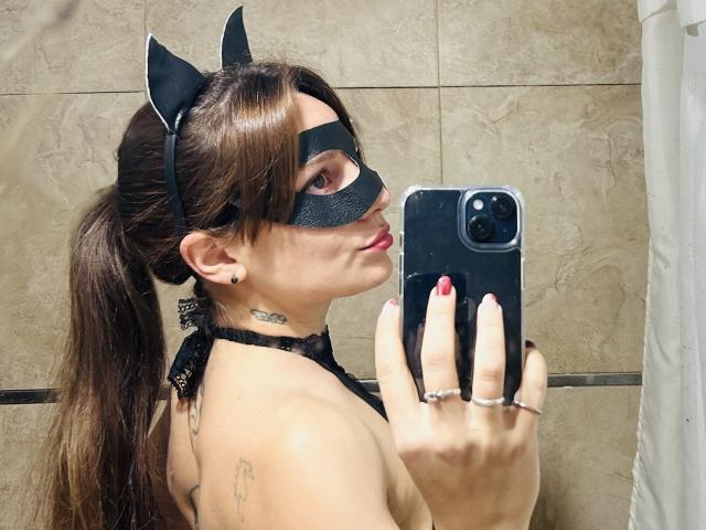 RomiSweety with brown hair in a ponytail, black cat mask, and lace halter, taking a mirror selfie with red nails, showing tattoos and a ring on her finger.