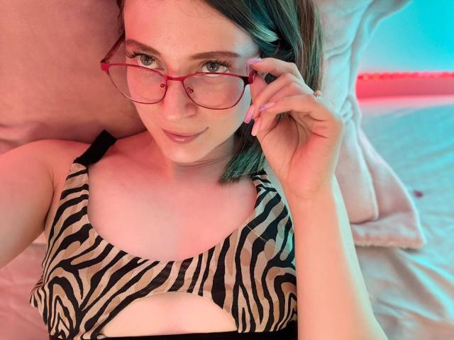 AmberWoody, con gli occhiali rossi, reggiseno a righe zebrate e un sorriso carino, tiene un bicchiere con la mano destra, sdraiata sul letto, con i capelli castani corti, illuminazione rosa-blu, che mostra i suoi piccoli seni..