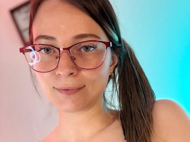 AmberWoody, com óculos vermelhos, rabo de cavalo castanho e pele clara, exibindo um sorriso discreto, com nariz pequeno e sardas, vestindo uma blusa preta com alças, com uma aparência adorável..