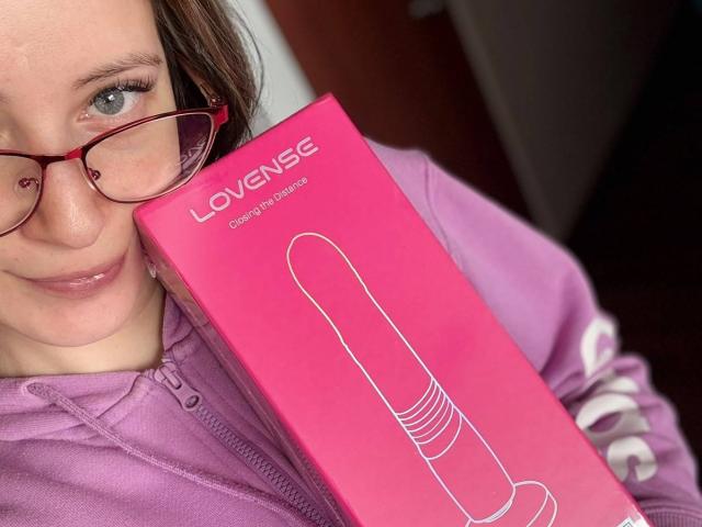 AmberWoody with red glasses holding a pink box of Loveise Realistic Dildo, wearing a purple hoodie, showing a playful smile and holding the dildo box against her cheek.