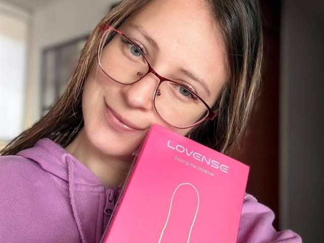 AmberWoody with red glasses, holding a pink LoveSense chocolate box, smiling softly, wearing a purple hoodie, her brown hair slightly messy, in a well-lit indoor setting.
