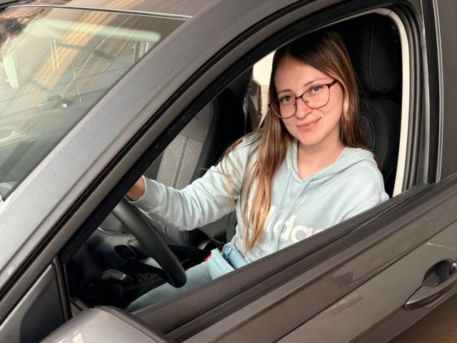 AmberWoody, de óculos, sentada em um carro cinza, vestindo um moletom azul claro, com as mãos no volante, sorrindo levemente, cabelos castanhos longos, lado direito do carro aberto..
