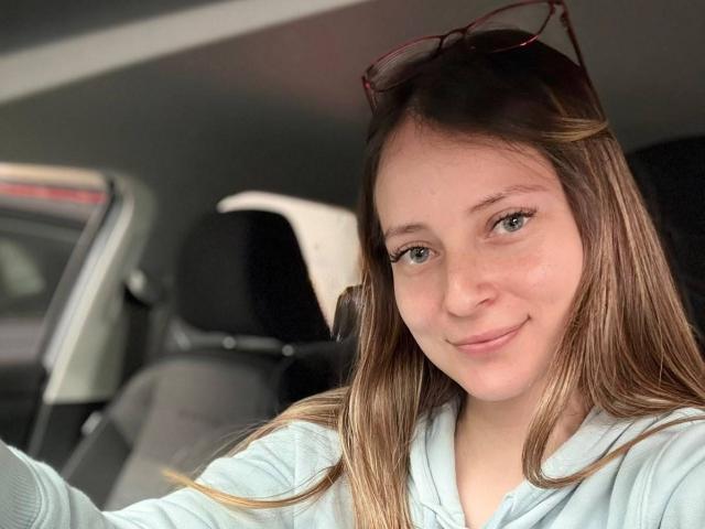 AmberWoody, com cabelo castanho claro, olhos azuis e um leve sorriso, vestindo um moletom azul claro, sentada em um carro com óculos de sol vermelhos na cabeça..