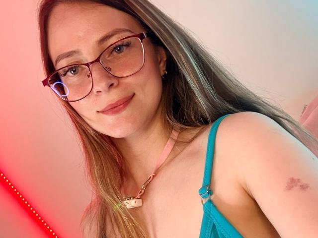 AmberWoody with long brown hair, glasses, pink necklace, blue bra, slight bruise on right shoulder, smiling softly, red light in background.