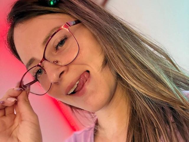 AmberWoody with glasses, tongue out, holding frame, brown hair with green hair clip, fair skin, small nose, light makeup, pink and blue light background, smiling, looking at camera.