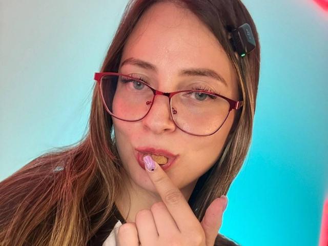 AmberWoody with red glasses, brown hair, and a headphone in her hair, sucking on a finger with a pink and yellow nail. Her nose has a septum piercing.