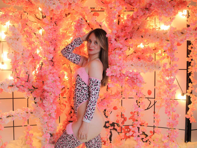 AmberWoody with pink and white blossoms, wearing a leopard print bodysuit, has a cute smile, hand in hair, showing her bare ass, in a pink-lit, floral setting.