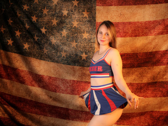 AmberWoody in een blauw en rood cheerleaderkostuum, die haar borsten en billen laat zien, staat met een glimlach, met bruin haar, voor een grote, gestructureerde Amerikaanse vlag..