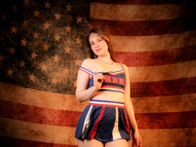 AmberWoody, con senos pequeños, vestida con un uniforme de animadora azul y rojo, sosteniendo una de las correas, sonriendo, de pie frente a una gran bandera estadounidense desgastada..