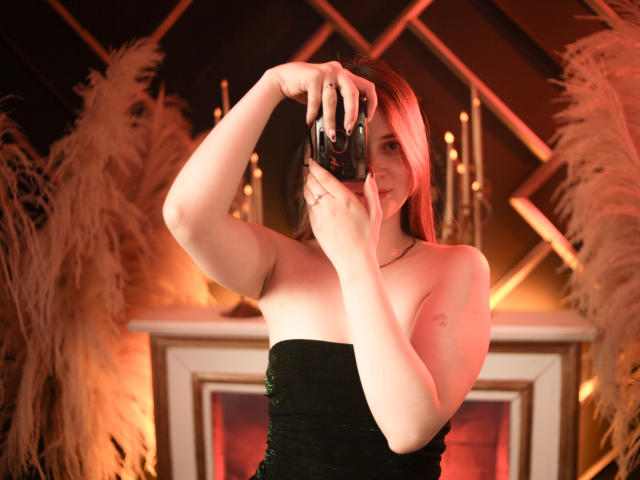 AmberWoody, con i capelli rossi, che tiene una macchina fotografica, sta scattando un selfie, indossa un abito nero senza spalline, mostrando le spalle scoperte e piccoli seni, con una macchina fotografica in mano..