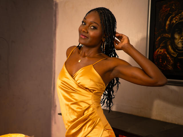 AndreaStorm with dark skin, long braids, and a yellow satin dress, showing her ass with one arm up, smiling at the camera. She's wearing a gold necklace.