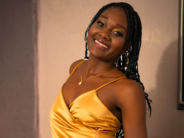 AndreaStorm with dark skin, smiling, wearing a yellow satin dress, braids, gold earrings, and a necklace, has a cute face and medium-sized tits.