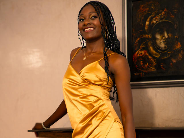 AndreaStorm with dark skin, long braided hair, smiling, wearing a shiny yellow satin dress, standing with hands on a wooden surface, showing her tits and ass.