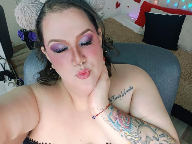 GiiannaMorrys con ombretto viola, guance rosa, piercing al naso, labbra carnose, tatuaggi sul braccio, mano al collo, una sedia grigia, capelli castani e un braccialetto rosso, che mette in mostra il suo seno..