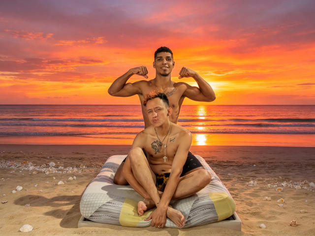 SantiagoAndLuke, muscular, shirtless, flexing, standing behind, has small tattoo on chest; Luke, shirtless, sits cross-legged on beach pillow, wearing black shorts, sunset, ocean, sandy shore.