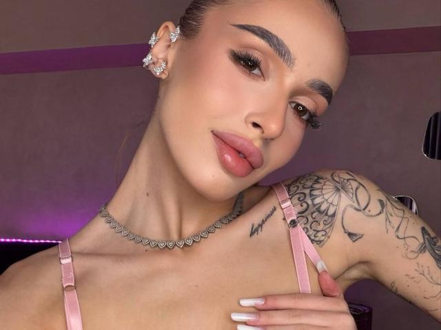 MarissaFex with pouty pink lips, brown eyes, and tattooed arm, wearing pink straps, heart necklace, and diamond earrings, holding her chest with white nails.