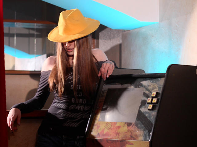 MiaNeit, com longos cabelos castanhos, usando um chapéu de cowboy amarelo, uma camisa preta com um ombro descoberto e jeans escuros, está apoiada em uma máquina de fliperama antiga com iluminação azul e branca..