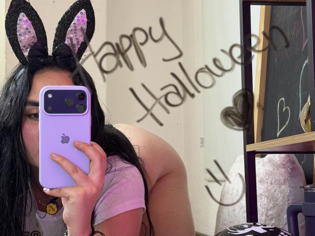 FenixAlthea, avec des cheveux noirs, portant une coque de téléphone violette, un haut violet, des oreilles de lapin noires, prenant un selfie, montrant ses fesses nues, tenant le téléphone devant son visage, fond blanc..