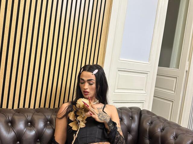 IrisIrs with black hair, pink hairclip, tattoos, eating a pastry on a brown tufted leather couch, wearing a black dress, with gold leaves in hand.