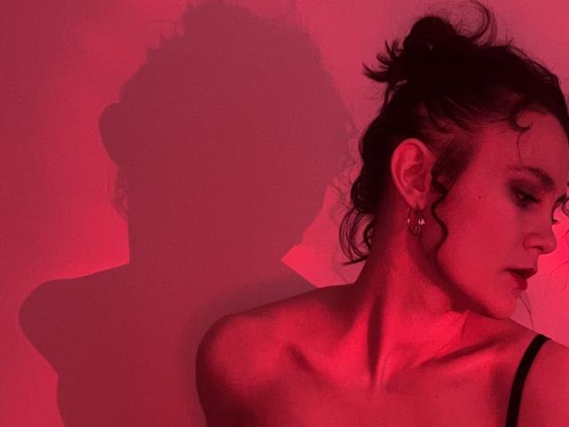 LunaNoirX with dark hair in a messy bun, wearing a black strap, red lit, shadow of her head and shoulders on the wall, small earrings, profile view.